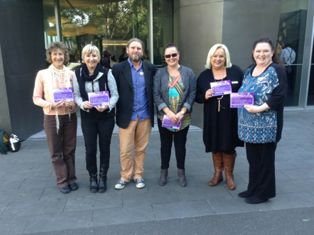  6 members from the Glenelg Transition Action Network standing in a row in front of a building, smiling and holding their awards from the Victorian Disability Sector Awards.